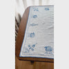 Video of off-white cross-stitch table topper spread across a dark wood table, with blue embroidered deer, trees and geometric motifs, finished with blue crocheted edging.