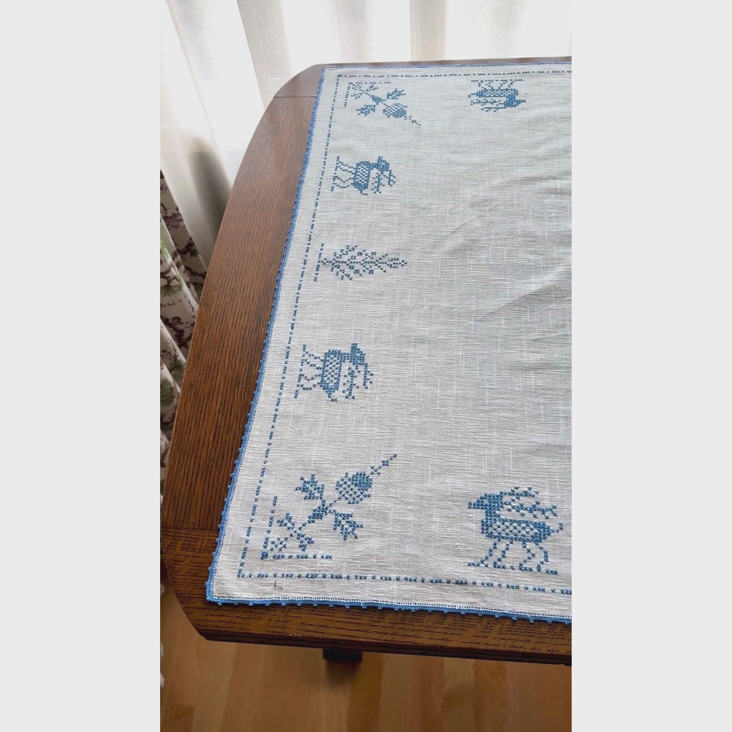 Video of off-white cross-stitch table topper spread across a dark wood table, with blue embroidered deer, trees and geometric motifs, finished with blue crocheted edging.