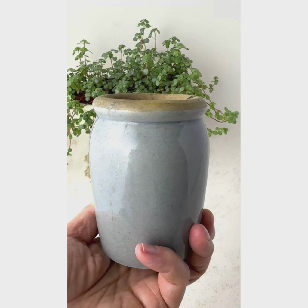 Hand holding small vintage gray-blue stoneware crock showing thick buff-colored rim, glazed exterior, and simple utilitarian form.