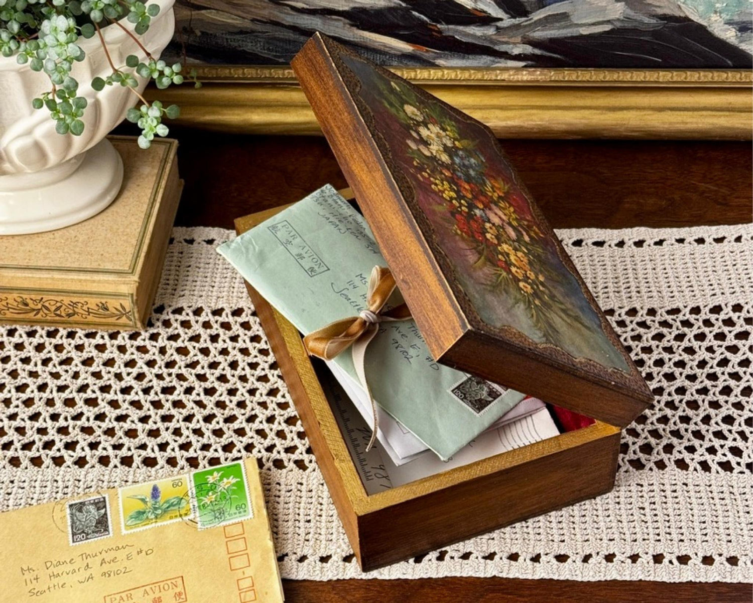 A vintage floral keepsake box resting open on a crocheted runner with handwritten letters tied in ribbon, vintage envelopes and postage stamps, beside a classical planter and framed artwork